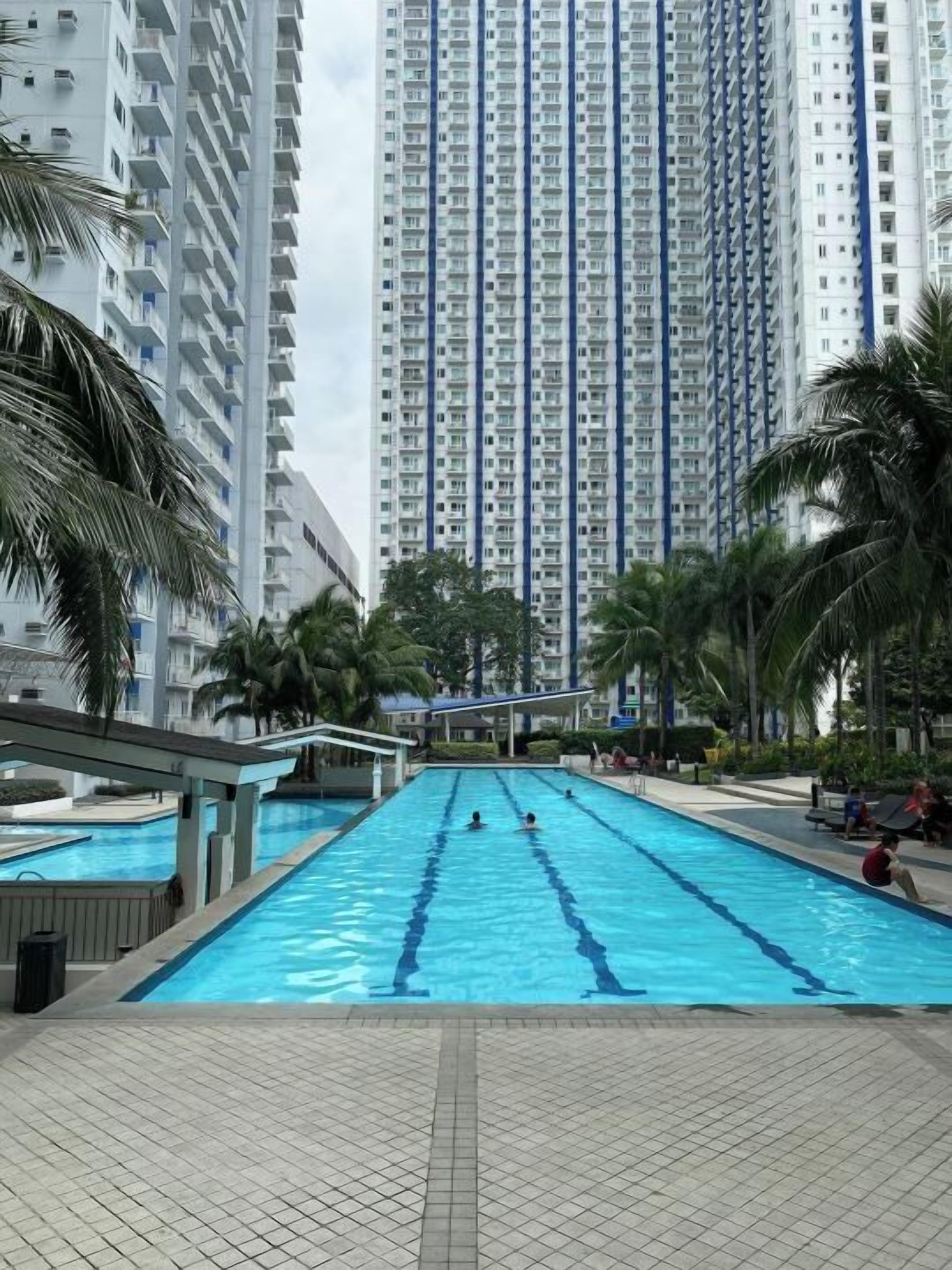A swimming pool with clear blue water, surrounded by palm trees and high-rise buildings in the background.