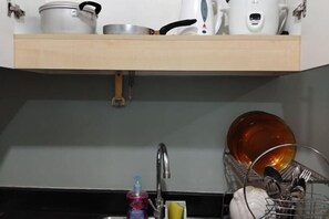 Cozinha com prateleira de madeira com panelas, chaleira e cafeteira. Abaixo da prateleira, pia com escorredor de louça contendo pratos e utensílios.