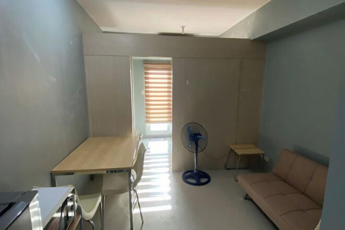 A small, well-lit room with a wooden desk, a chair, a standing fan, a sofa, and a window with blinds.