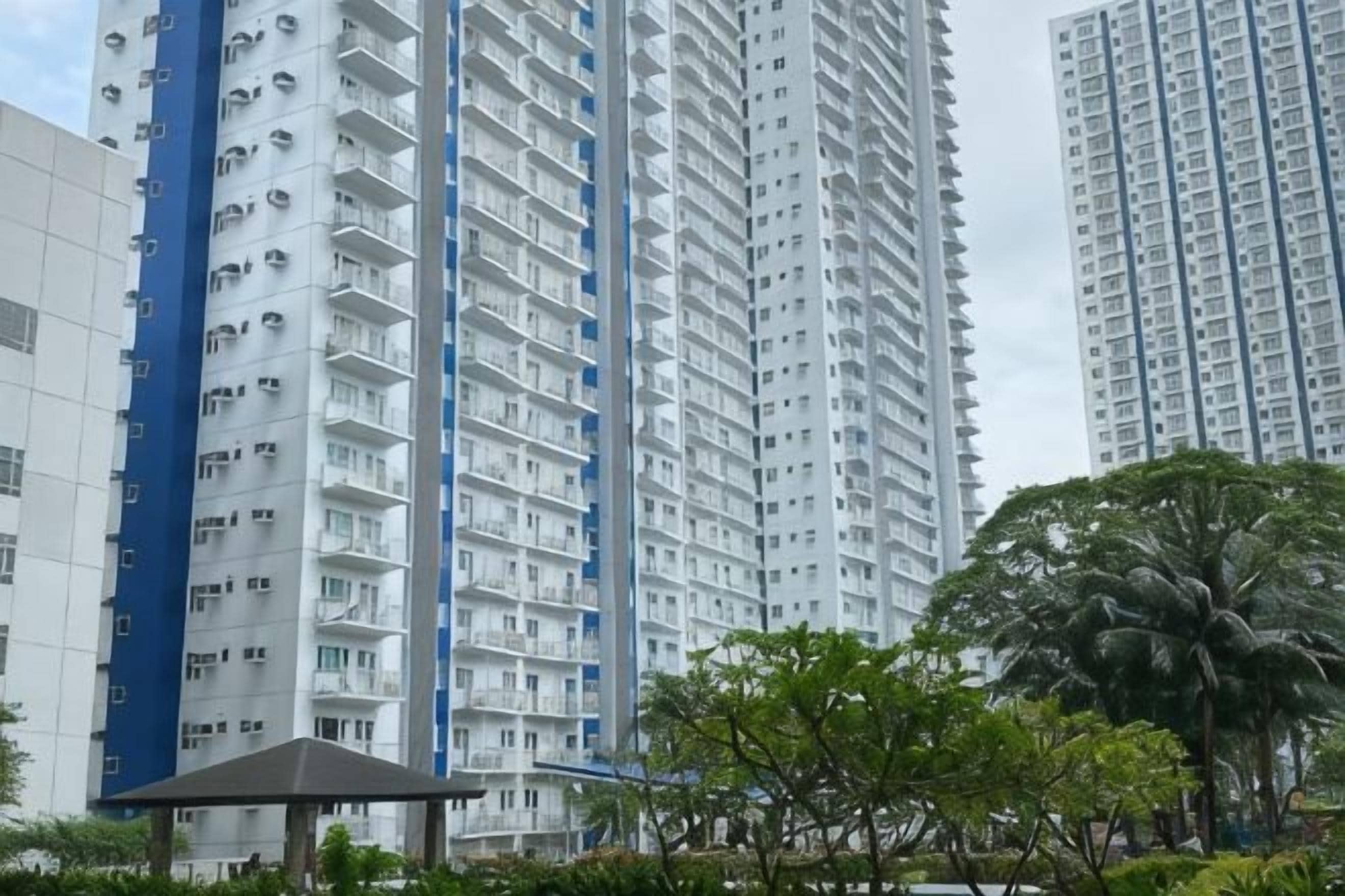 A high-rise residential building with multiple balconies, surrounded by greenery and a small pavilion.