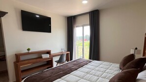 A hotel room with a bed, a desk, a chair, a TV mounted on the wall, and a view of the outdoors through a window. - Quinta Candela Hotel (Chignahuapan)