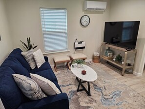 A living room with a blue sofa, a white coffee table, a TV on a wooden stand, a clock on the wall, and a window with blinds.