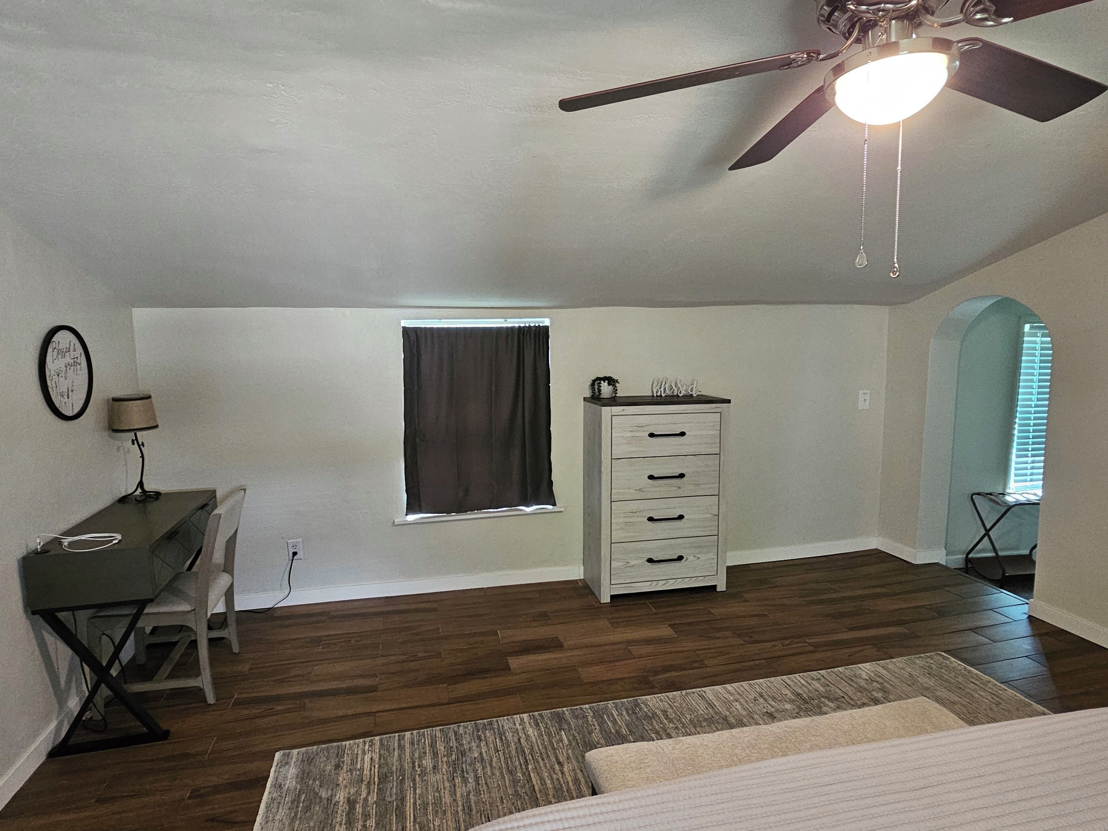 Una habitación con ventilador de techo, un escritorio con lámpara, cómoda, ventana con cortinas y suelo de madera.