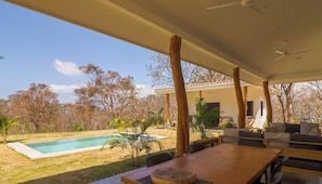 A covered outdoor area with a pool, wooden furniture, and a view of the surrounding landscape.
