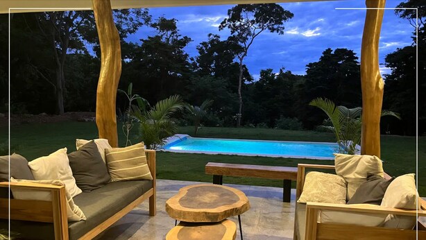 An outdoor seating area with a pool view, featuring wooden furniture and a stone table.