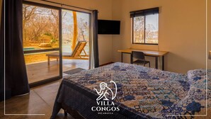 A bedroom with a large bed, a desk, and a TV. There is a sliding glass door leading to an outdoor area with a chair and a table.