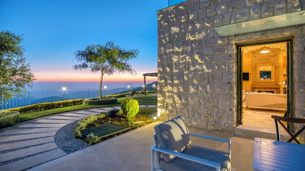 A stone building with a glass entrance, outdoor seating, and a view of the sea at dusk. - Red Oak House With Garden & Terrace in Assia, Batroun (Bchaaleh)
