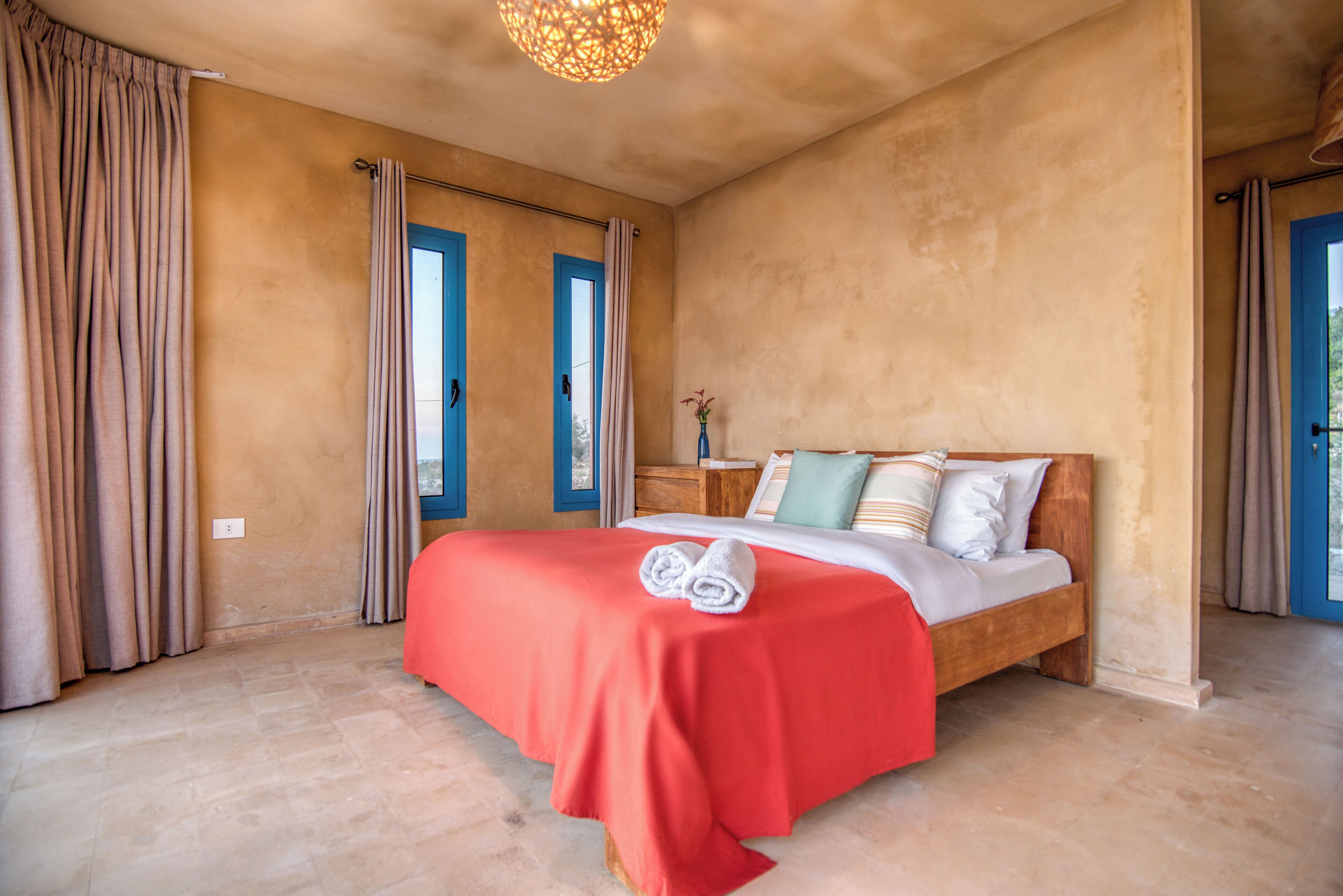 A bedroom with a wooden bed, beige curtains, and a red bedspread.