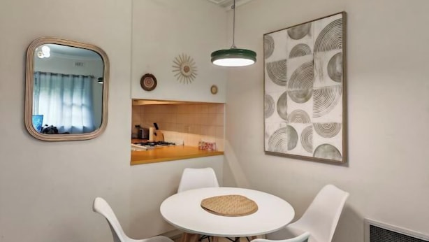 A dining area with a round table, white chairs, a hanging pendant light, a mirror, and a wall-mounted artwork.