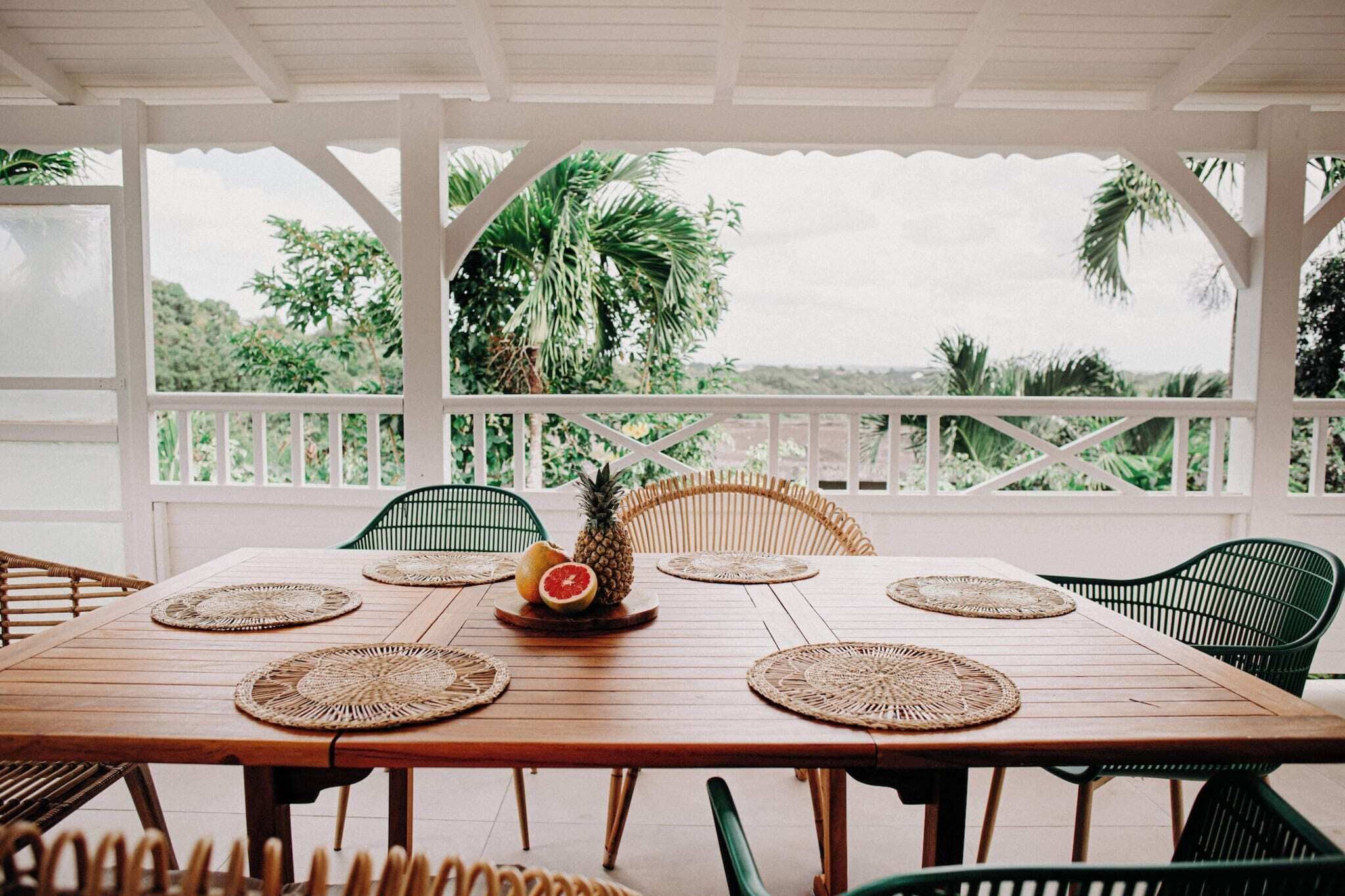 A wooden table with woven placemats, surrounded by wicker chairs, set in a covered outdoor area with a view of lush greenery.