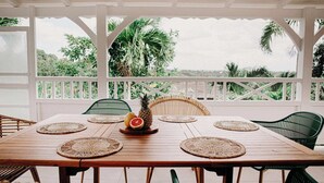 Une table en bois, agrémentée de sets de table en fibres tressées, entourée de chaises en osier, installée dans un espace extérieur abrité offrant une vue sur une végétation luxuriante.