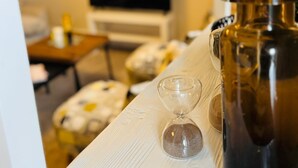 Ett rum med ett vitt träbord, en mörk kanna och ett genomskinligt glas. I bakgrunden står en tv på ett vitt stativ och en soffa med blommiga kuddar.
