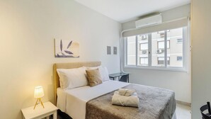Un dormitorio ordenado con cama, lámpara de noche y ventana con vista a edificios.