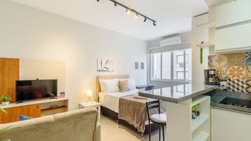 Quarto de hotel moderno com uma cama, TV, escrivaninha e cozinha compacta.