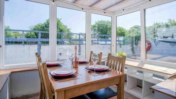 A dining area with a wooden table set for four, large windows, and a view of greenery and a fence.