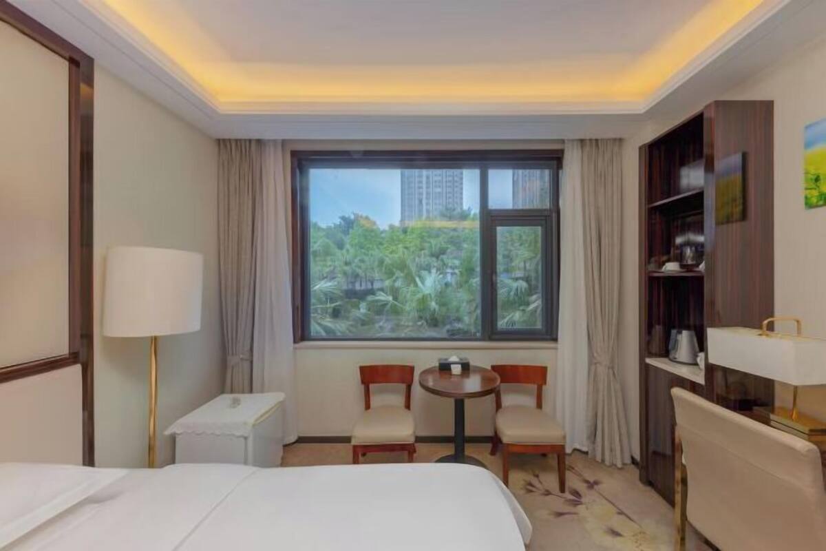 A hotel room with a bed, a small table with two chairs, a window with curtains, and a bookshelf.