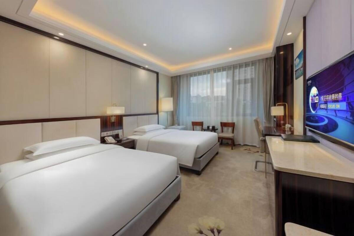 A hotel room with two beds, a desk, a TV, and a window with curtains.
