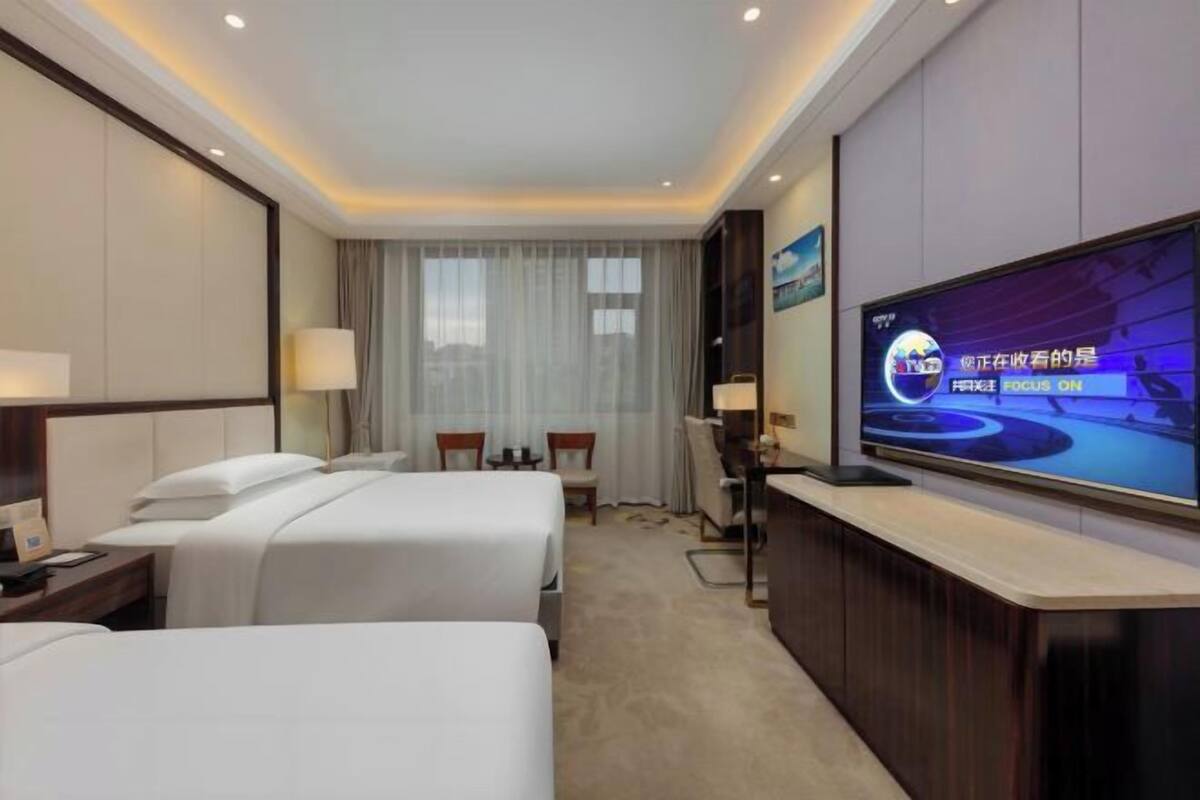 A hotel room with two beds, a desk, a TV, and a large window with curtains.