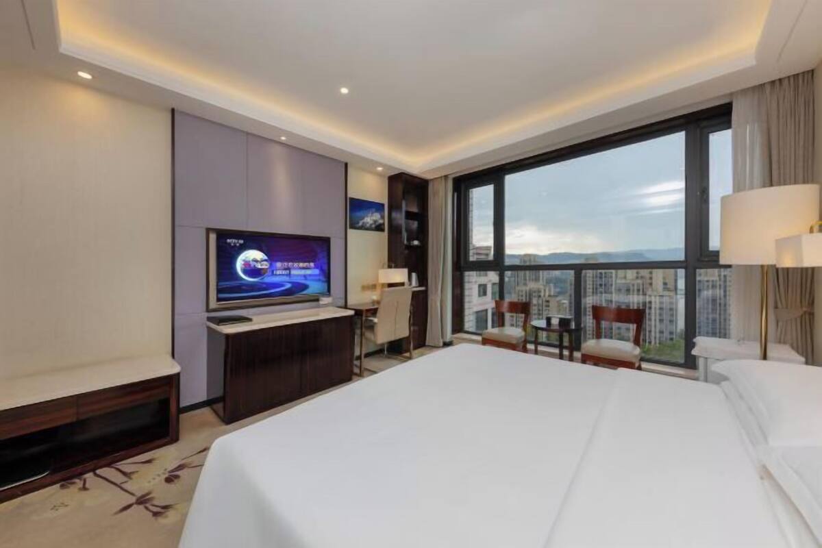 A hotel room with a large bed, a TV, a desk, and a view of the city.