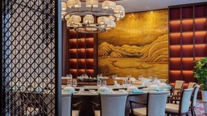 Un'area pranzo con un tavolo rotondo apparecchiato per sei persone, una parete decorata con un paesaggio dorato e un lampadario moderno.