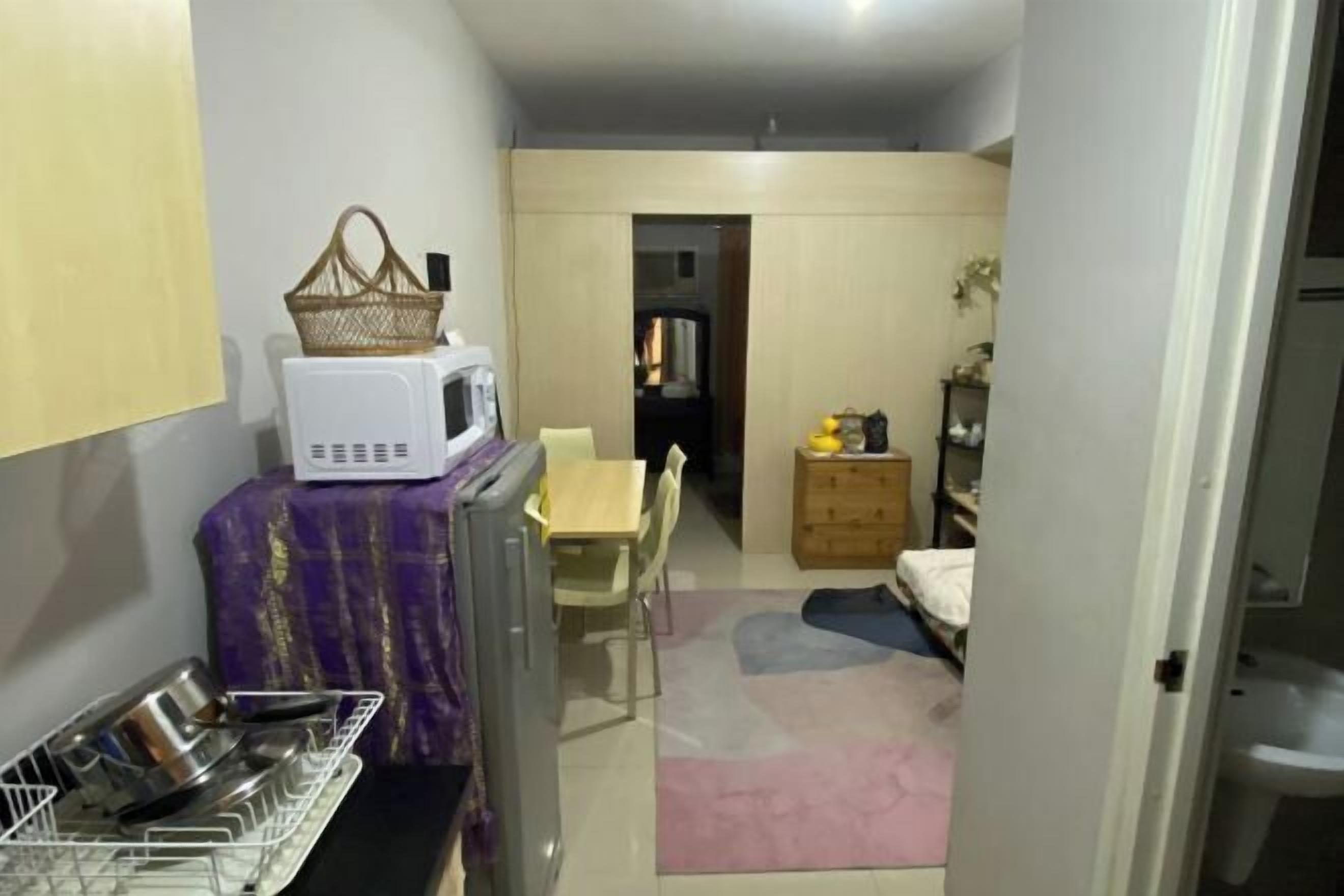A compact kitchen with a microwave, a basket, and a dish rack. There is a dining area with a table and chairs, and a bed in the background.