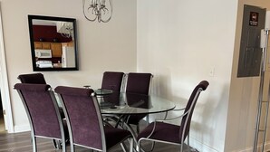 Een eetruimte met een ronde glazen tafel, zes paarse gestoffeerde stoelen, een kroonluchter en een magnetron op de achtergrond.