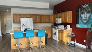 Een moderne keuken met houten kasten, een witte koelkast, een magnetron en een fornuis. Drie blauwe barkrukken en een klein eiland met een spoelbak.