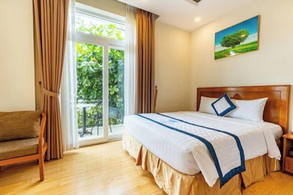 Une chambre d’hôtel avec un lit, une chaise en bois, une table de chevet et une vue sur la verdure, visible à travers une porte vitrée.