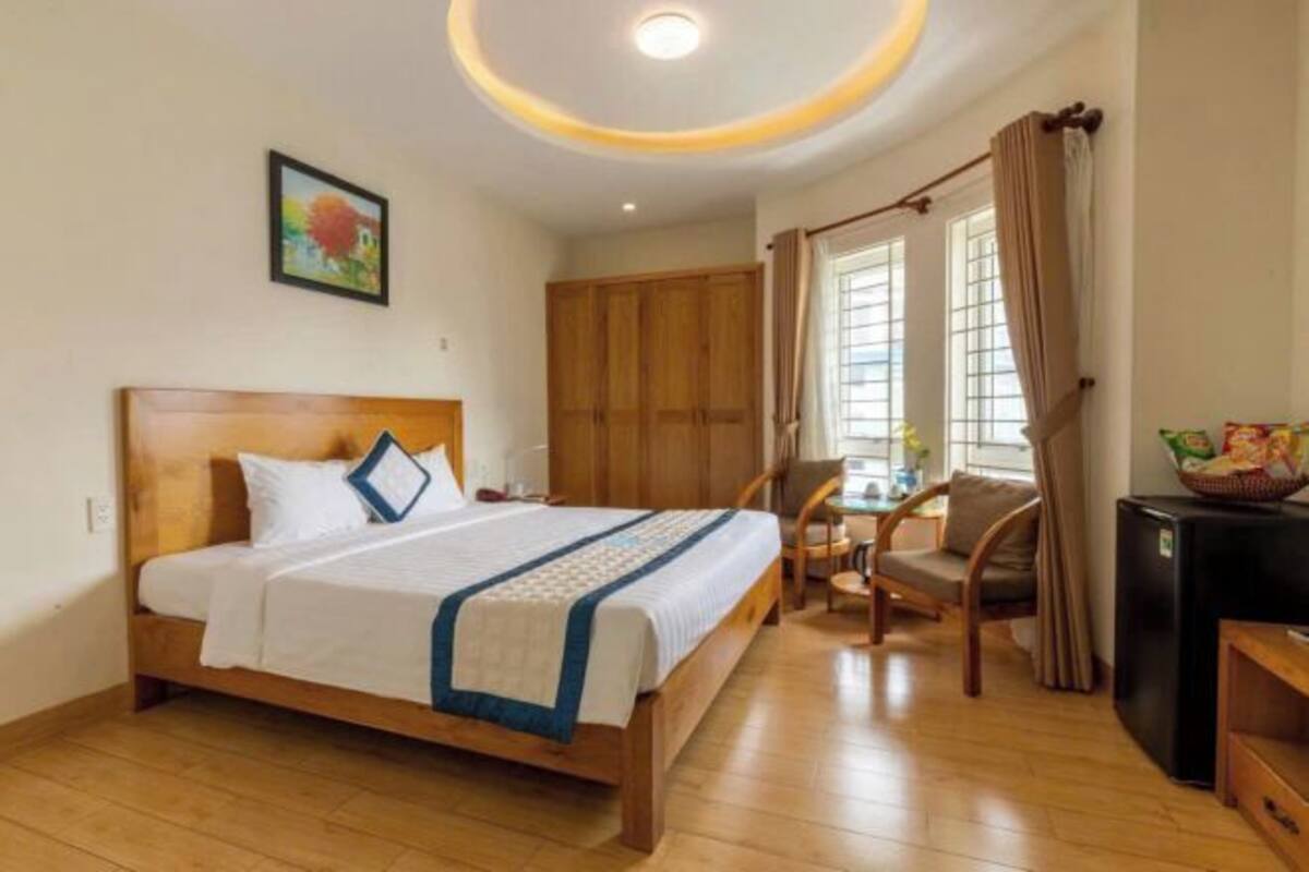 Une chambre d’hôtel avec un lit en bois, un bureau, une chaise, une petite table et une fenêtre avec des rideaux.