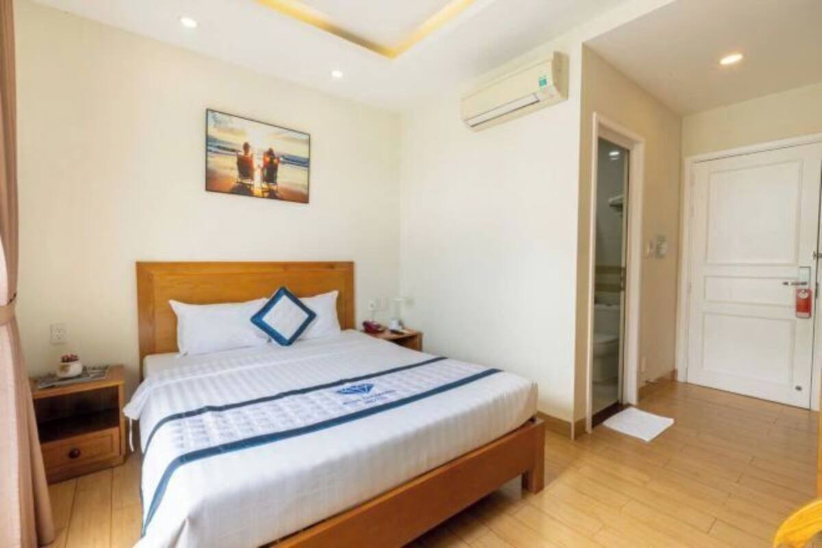 Une chambre d’hôtel avec un lit en bois, une table de chevet, un climatiseur fixé au mur, une porte et un tableau encadré accroché au mur.