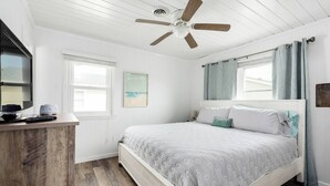 Ein Schlafzimmer mit einem Bett, einem Deckenventilator, einem Fenster und einem Holztisch.