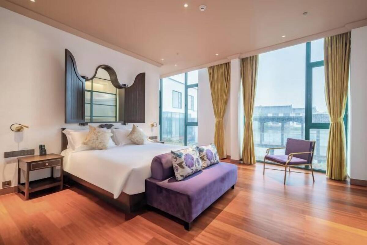 A modern hotel room with a large bed, a bench, a chair, and a large window with curtains.