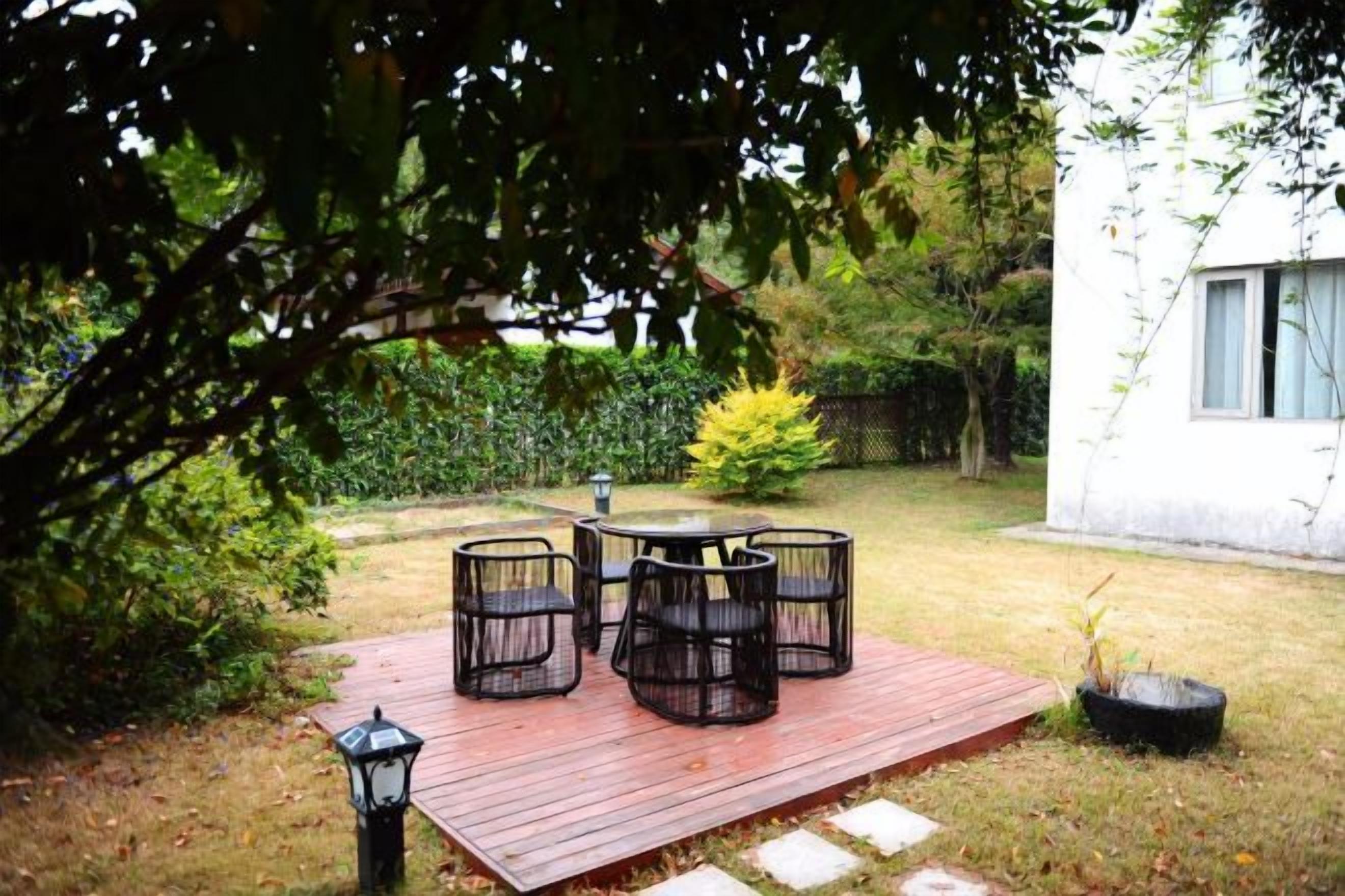 A patio with a table and chairs, surrounded by a wooden deck, in a garden with a white building in the background.
