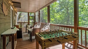 Une terrasse en bois avec une table de baby-foot, des meubles en osier et de grandes fenêtres donnant sur une forêt.