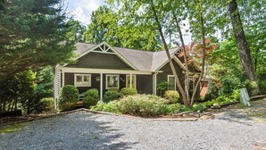 Une maison de plain-pied avec un toit en pente, entourée d’arbres denses et bordée d’une allée en gravier.