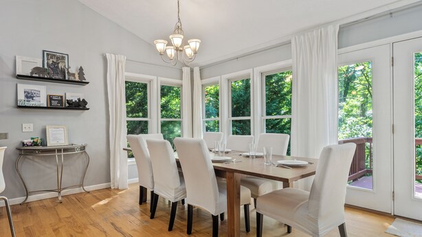 Un coin repas avec une table en bois, des chaises blanches et un lustre. Il y a des fenêtres avec des rideaux blancs, et une petite table ornée d’objets décoratifs est fixée au mur.