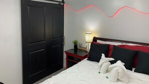 A bedroom with a black sliding door, a bed with white bedding and red accents, a bedside table with a lamp, and a wall-mounted light fixture.
