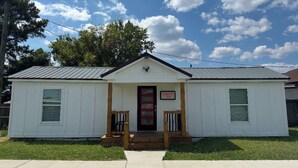 A small, single-story building with a white exterior and a red door. It has a small porch with wooden railings and steps leading up to the door.