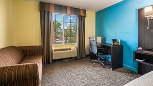 Une chambre d’hôtel avec un canapé à rayures, un bureau avec une chaise, une fenêtre avec des stores, une lampe fixée au mur et une enseigne indiquant « Magnuson ».
