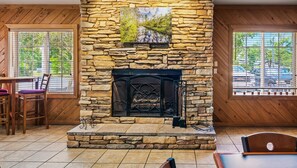 A stone fireplace with a painting above it, wooden walls, a dining area with chairs, and a view of parked cars through large windows. - Relax in Style: King Bed Non-Smoking Room at Wildwood Inn (Crawfordville)