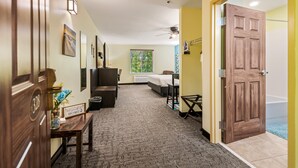 Una habitación de hotel con cama, escritorio, silla, mesita de noche con un florero, un cubo de basura y una puerta de madera.