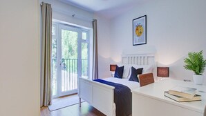 A bedroom with a bed, bedside tables, a desk, a potted plant, and a window with curtains. - Hybridresi New Malden (New Malden)