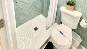 A bathroom with a herringbone tile pattern, a shower with a handheld showerhead, a toilet with a plant on top, and a small shelf with toiletries.