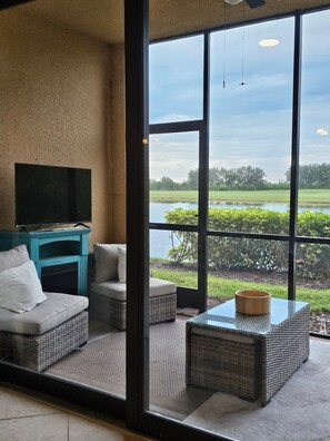 A room with a large window offering a view of a lake and greenery. There is a sofa, a small table, and a television on a stand. - 2br/ 2bth spacious garden condo perfect for golfer w deeded golf course access (Bradenton)
