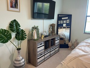 Une chambre à coucher avec un lit, un téléviseur fixé au mur, une commode avec des tiroirs, une grande plante et un mur orné de tableaux.
