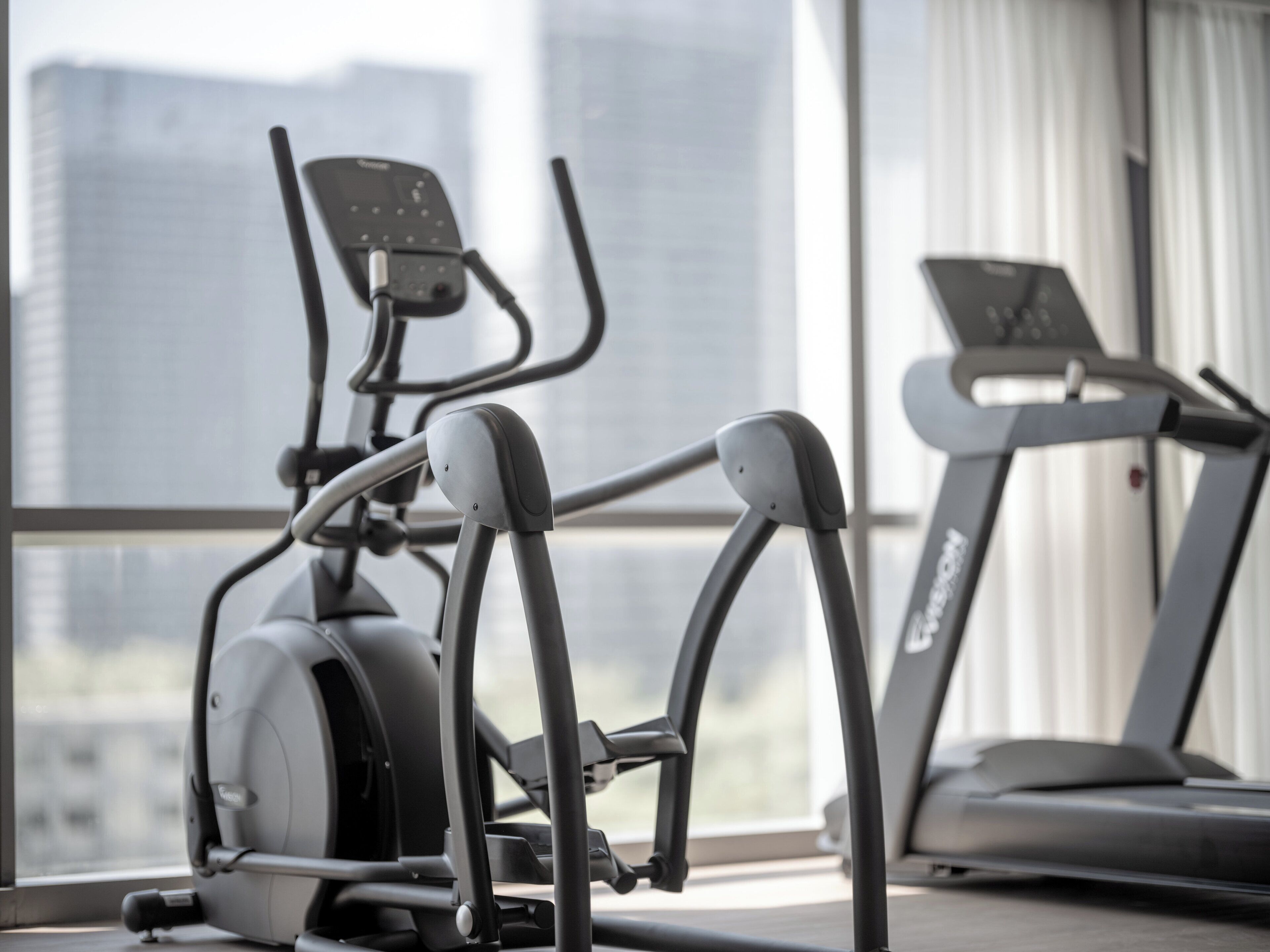An indoor gym with exercise equipment, including an elliptical and a treadmill, with a city view through large windows.