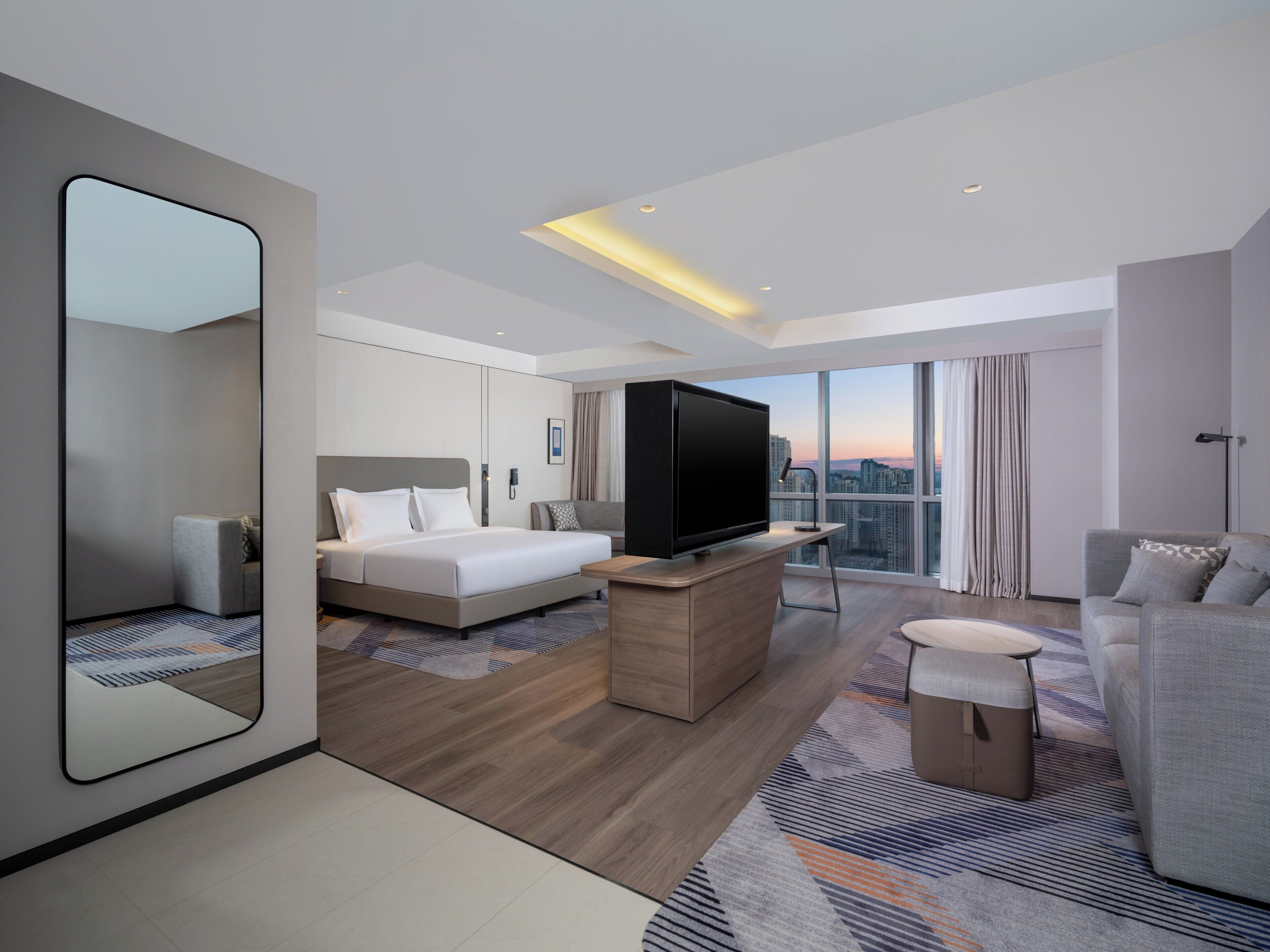 Una habitación de hotel moderna con una cama grande, un televisor sobre un soporte de madera, un sofá, una mesita pequeña y un ventanal con vistas a la ciudad.