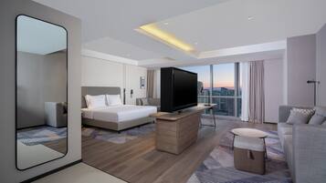 Una habitación de hotel moderna con una cama grande, un televisor sobre un soporte de madera, un sofá, una mesita pequeña y un ventanal con vistas a la ciudad.
