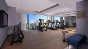 Une salle de sport moderne équipée de tapis de course, de vélos elliptiques et d’un rameur. Elle dispose de grandes fenêtres offrant une vue sur la ville et d’un banc bleu.