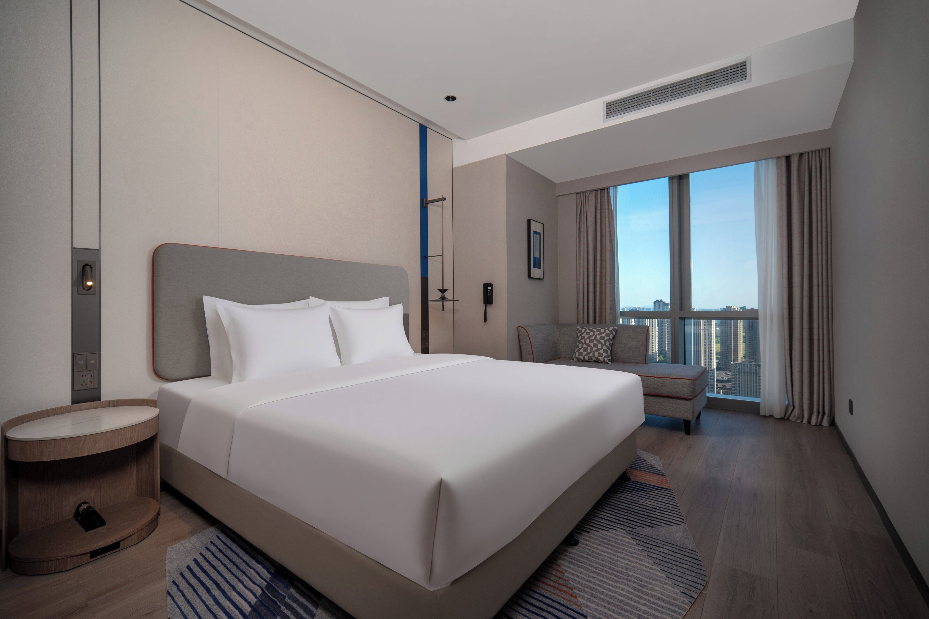 Une chambre d’hôtel moderne dotée d’un grand lit, d’une table de chevet, d’un petit coin salon et offrant une vue sur la ville grâce à de grandes fenêtres.